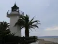 El faro de Palamós, blanco y esbelto, se alza junto a palmeras y el mar azul.