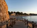 Pasarela de madera hacia la platja Sant Pol, Sant Feliu de Guíxols. Edificios blancos y palmeras bordean la arena dorada bajo un cielo azul.