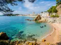Una caleta en la Costa Brava con aguas cristalinas, buceadores explorando el fondo rocoso y una playa con gente disfrutando del sol.