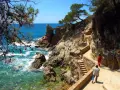 Un camino de ronda con escaleras de piedra serpentea por la costa rocosa. El mar turquesa rompe contra las rocas.