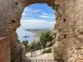Vista desde el **castillo de Blanes** hacia la bahía y la ciudad. El mar azul contrasta con las ruinas de piedra y el cielo nublado.