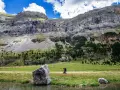 Senderista camina por un prado verde con imponentes montañas de **3000 del Pirineo** al fondo.