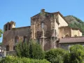 El **monasterio de Siresa**, con su imponente arquitectura de piedra y campanario, se alza bajo un cielo azul, rodeado de vegetación exuberante.