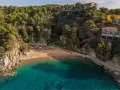 Bahía de Cala Bona, Barcelona, con aguas turquesas, arena dorada y acantilados verdes. Casas en la ladera.