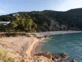 La platja de Canyerets, con su arena dorada y aguas cristalinas, se extiende bajo una colina verde y casas blancas.