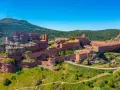 Imponente castillo de Peracense, una fortaleza de piedra rojiza que se alza sobre un peñón, rodeada de vegetación y bajo un cielo azul intenso.