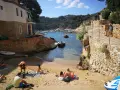 Gente relajándose en la arena de cala Malaret, Begur, con barcos anclados en aguas cristalinas y colinas verdes al fondo.