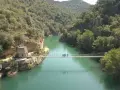 Un puente colgante cruza un río turquesa rodeado de montañas verdes. Un grupo de personas camina por el puente de Montañana.