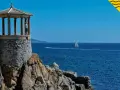 Un mirador de piedra con columnas se alza sobre rocas, con el mar azul y veleros al fondo. Un lugar perfecto para pasear por los **caminos de ronda**.