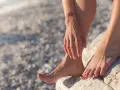 Pies descalzos sobre rocas en una playa nudista de Cadaqués. El sol brilla sobre la piel mojada y un pequeño tatuaje de flores adorna el brazo.