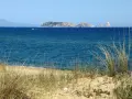Dunas de hierba seca en primer plano, mar azul y un archipiélago rocoso en la distancia, evocando la belleza de la playa mas Pinell.