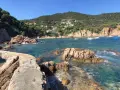 Un puente de piedra cruza aguas turquesas en la cala Canyet. Barcas flotan tranquilas mientras colinas verdes y casas blancas coronan el paisaje.
