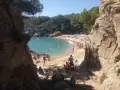 Gente disfrutando del sol y el mar en la platja de Treumal, con rocas y pinos en primer plano.