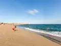 Un niño juega en la arena dorada de la playa de les barques, mientras otros disfrutan del mar azul y un velero navega en el horizonte.