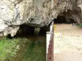 Entrada a una cueva con un arroyo y vegetación. Un camino pavimentado invita a explorar qué ver en Ribadesella.
