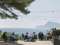 Gente disfrutando de una comida al aire libre con vistas al mar. Un lugar ideal donde comer en Altea, con mesas azules y un ambiente relajado bajo un árbol.