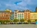 Fachada de edificios coloridos en una plaza, ideal para saber **qué ver en Pamplona**. Terrazas y árboles verdes bajo un cielo azul.
