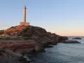 Faro sobre acantilados rojizos, con el mar azul y un cielo despejado. Un camino desciende hacia una pequeña cala roja.