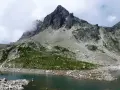 Paisaje montañoso con un lago de aguas tranquilas en primer plano. Las laderas están cubiertas de rocas y vegetación verde, con picos escarpados y nubes en el cielo. Los **ibones de Arriel** reflejan la majestuosidad del entorno.