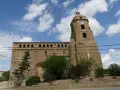 Iglesia de piedra con torre y reloj, rodeada de vegetación. Un día soleado en Alberuela de Tubo.