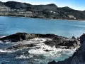 Olas rompiendo en rocas oscuras, con la costa verde y edificios de Mundaka al fondo. Un lugar perfecto para comer y disfrutar del paisaje.