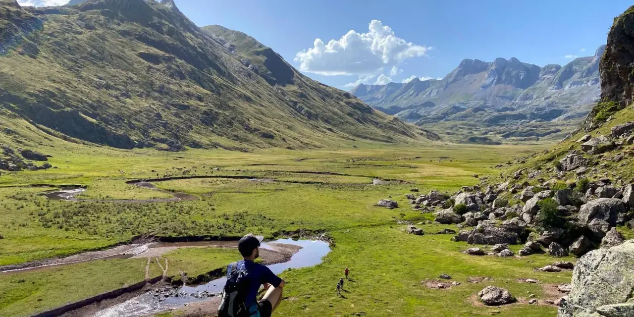 Un excursionista contempla un vasto valle de hecho, con un río serpenteante y montañas majestuosas bajo un cielo azul.
