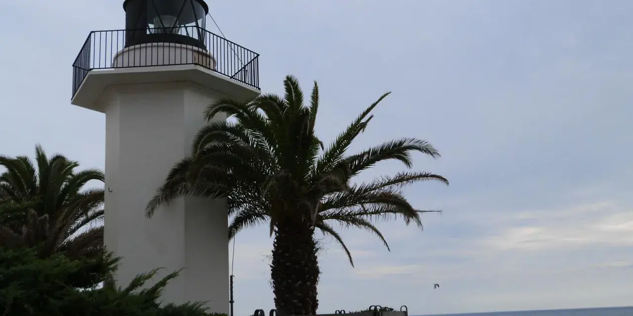 El faro de Palamós, blanco y esbelto, se alza junto a palmeras y el mar azul.