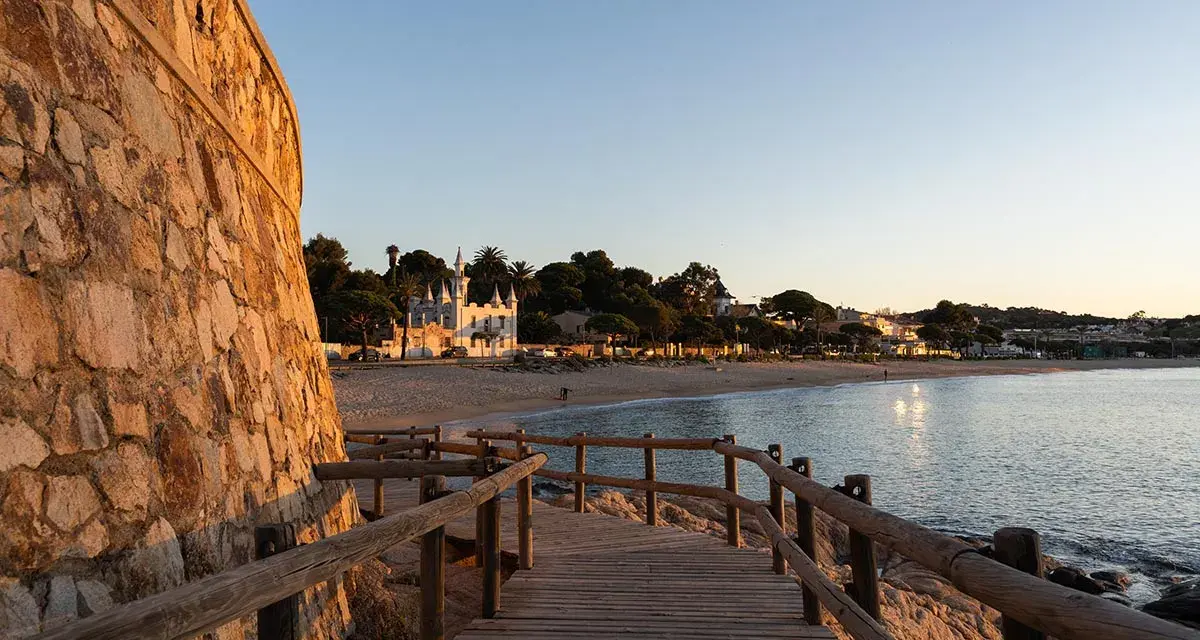 Pasarela de madera hacia la platja Sant Pol, Sant Feliu de Guíxols. Edificios blancos y palmeras bordean la arena dorada bajo un cielo azul.