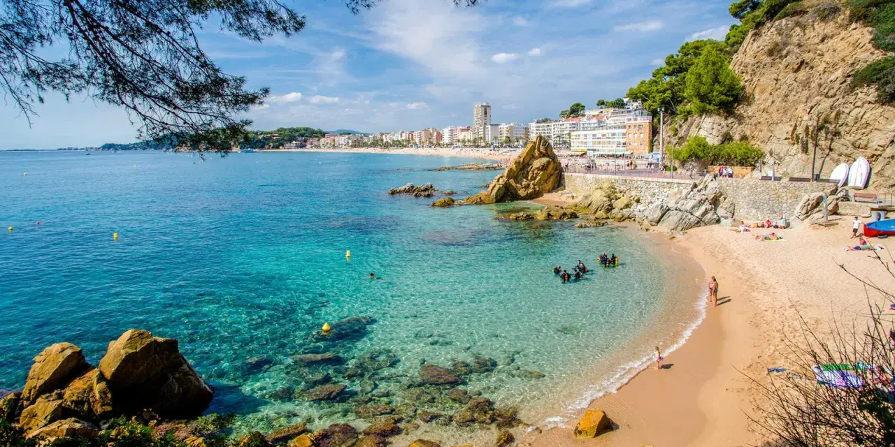Una caleta en la Costa Brava con aguas cristalinas, buceadores explorando el fondo rocoso y una playa con gente disfrutando del sol.