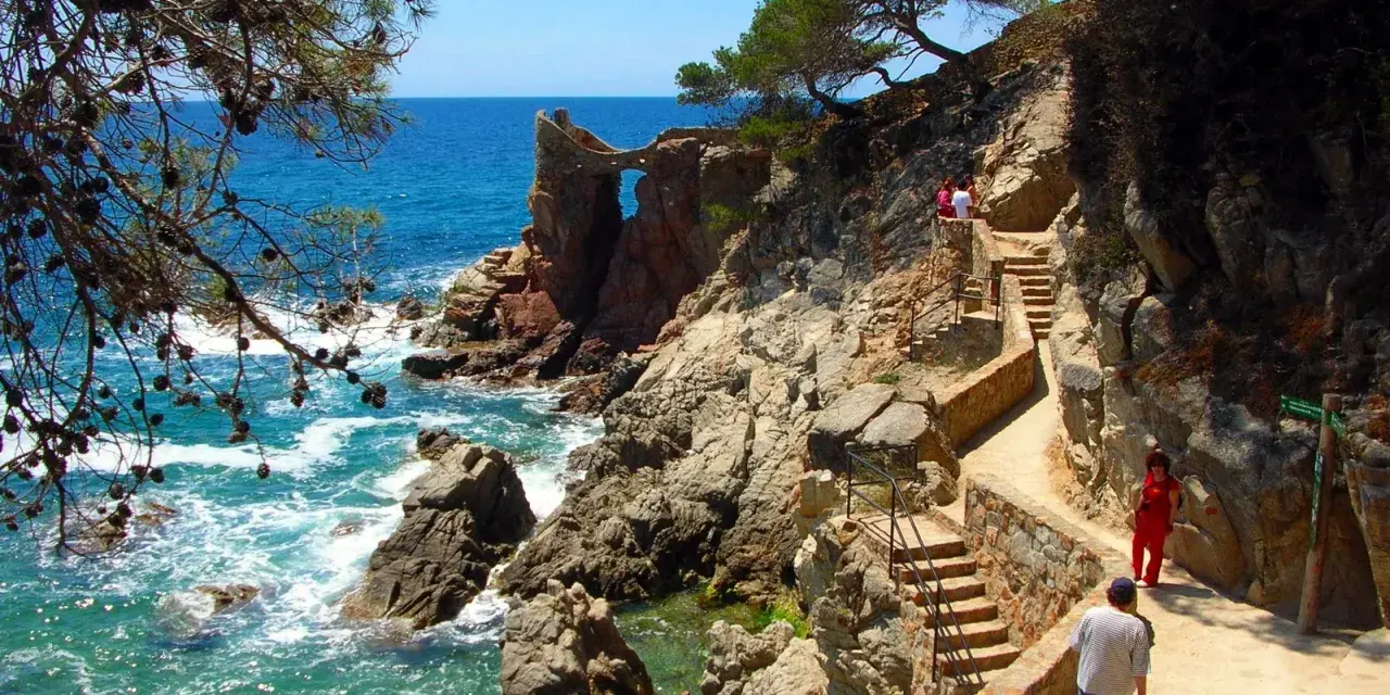 Un camino de ronda con escaleras de piedra serpentea por la costa rocosa. El mar turquesa rompe contra las rocas.