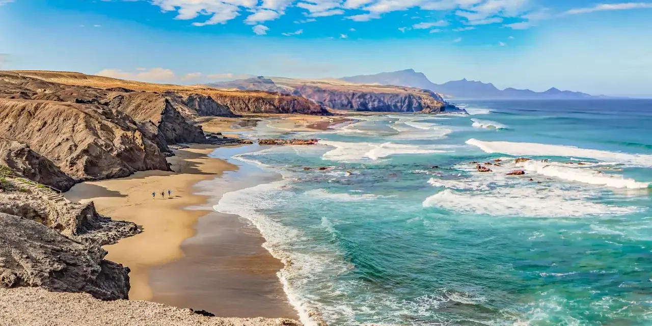 Paisaje de las playas de Fuerteventura con acantilados rocosos, arena dorada y el mar turquesa con olas rompiendo.