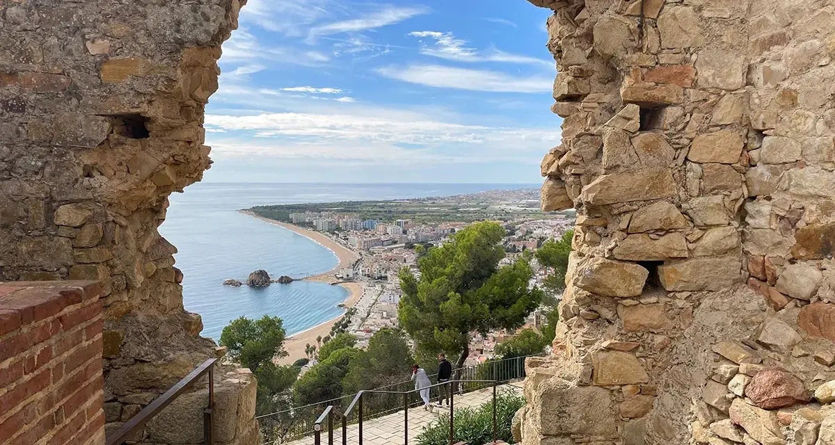 Vista desde el **castillo de Blanes** hacia la bahía y la ciudad. El mar azul contrasta con las ruinas de piedra y el cielo nublado.