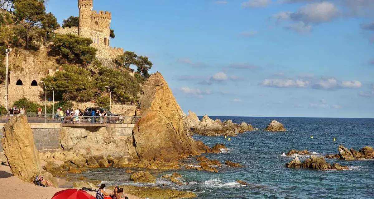 Castillo medieval sobre acantilados rocosos en sa caleta Lloret de Mar. Playa con gente y mar azul.