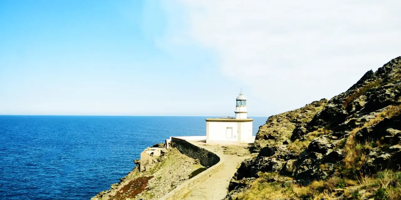 Un faro blanco se alza sobre un acantilado rocoso, vigilando el mar azul. Un camino serpentea hacia él, invitando a explorar los **faros Costa Brava**.
