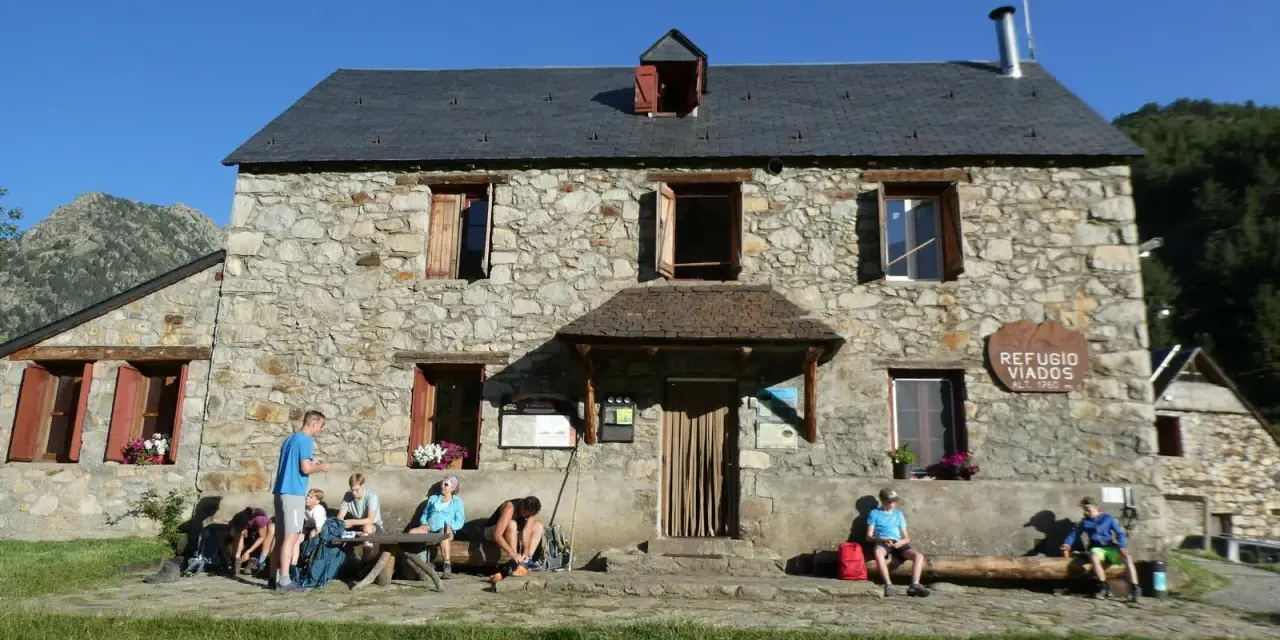 Grupo de excursionistas descansa frente al Refugio Viados, parte de la ruta de los tres refugios. Montañas y cielo azul al fondo.