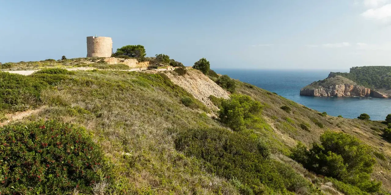 La torre de Montgó se alza sobre una colina verde, vigilando el mar azul y una bahía rocosa.