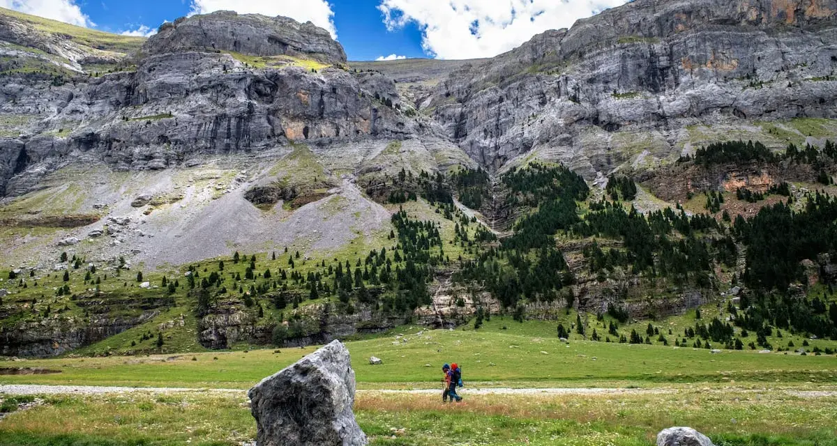 Senderista camina por un prado verde con imponentes montañas de **3000 del Pirineo** al fondo.