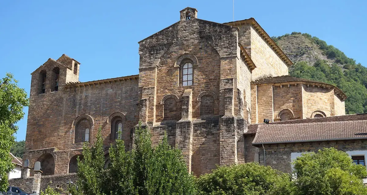 El **monasterio de Siresa**, con su imponente arquitectura de piedra y campanario, se alza bajo un cielo azul, rodeado de vegetación exuberante.