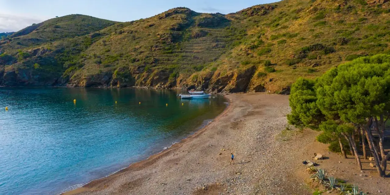Tranquila cala Joncols plage con aguas turquesas, una persona paseando y barcas amarradas.