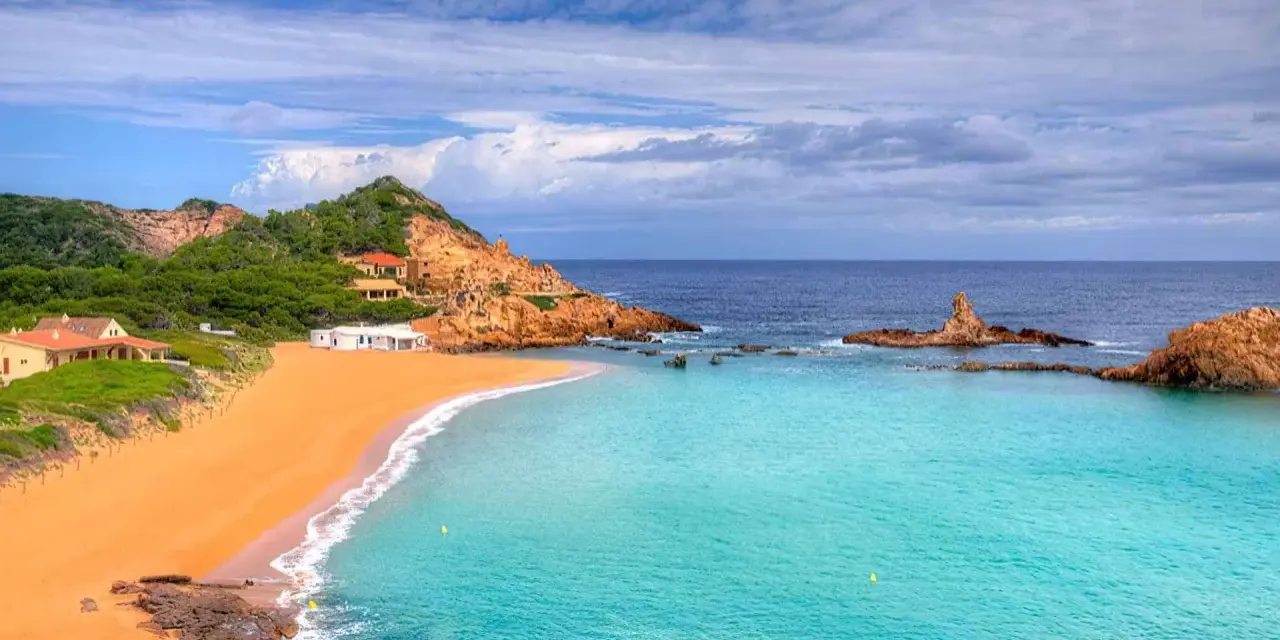 Hermosas calas cerca de Fornells, con arena dorada, aguas turquesas y casas blancas integradas en la costa rocosa.