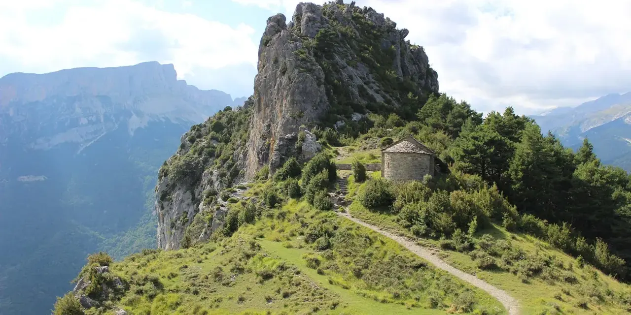 Pequeña ermita de piedra con tejado cónico, rodeada de vegetación y un sendero que sube por la ladera. Las ermitas de Tella se alzan majestuosas.