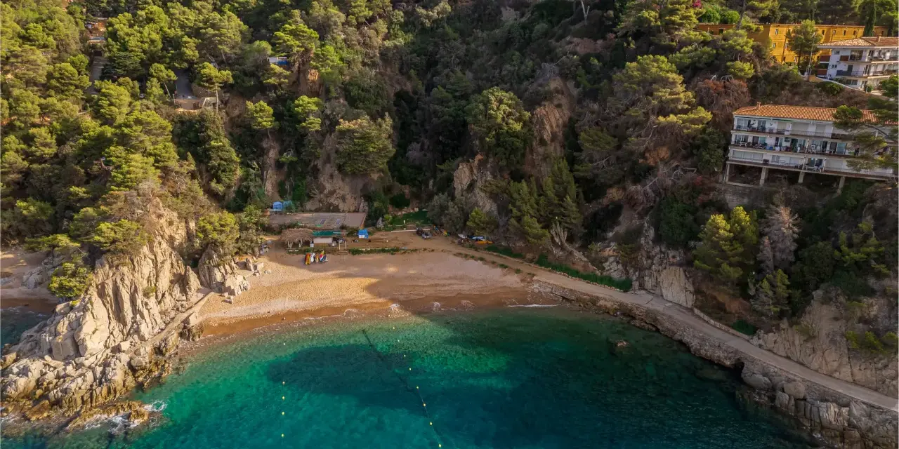 Bahía de Cala Bona, Barcelona, con aguas turquesas, arena dorada y acantilados verdes. Casas en la ladera.