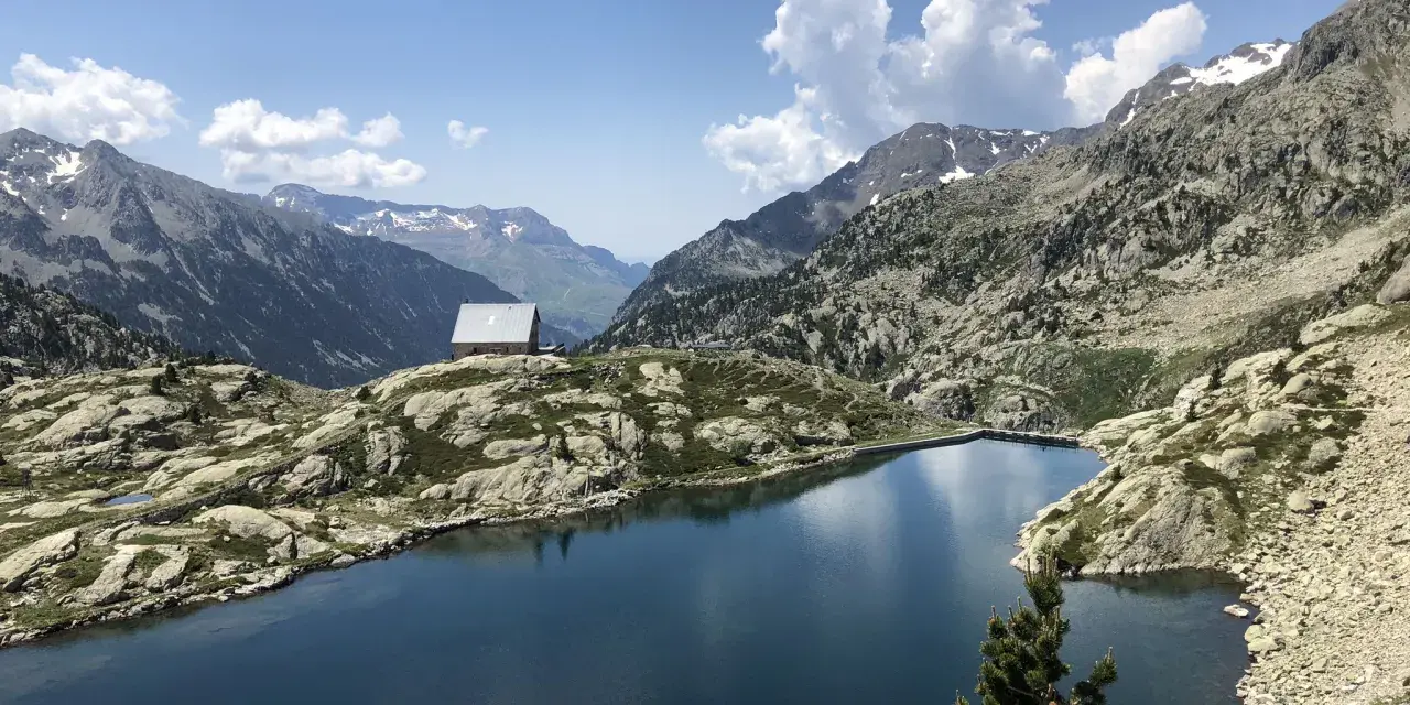 Un refugio solitario junto a los ibones de Bachimaña, rodeado de montañas rocosas y un lago azul intenso bajo un cielo despejado.