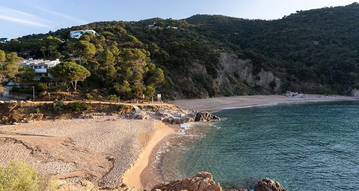 La platja de Canyerets, con su arena dorada y aguas cristalinas, se extiende bajo una colina verde y casas blancas.