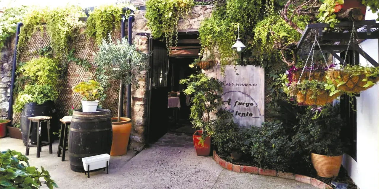 Entrada a un restaurante con vegetación exuberante. Un cartel dice "Restaurante A fuego lento". Ideal para saber donde comer en Hervás.
