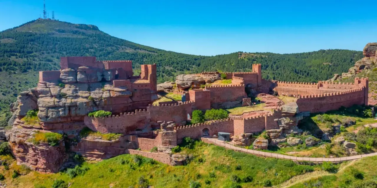 Imponente castillo de Peracense, una fortaleza de piedra rojiza que se alza sobre un peñón, rodeada de vegetación y bajo un cielo azul intenso.
