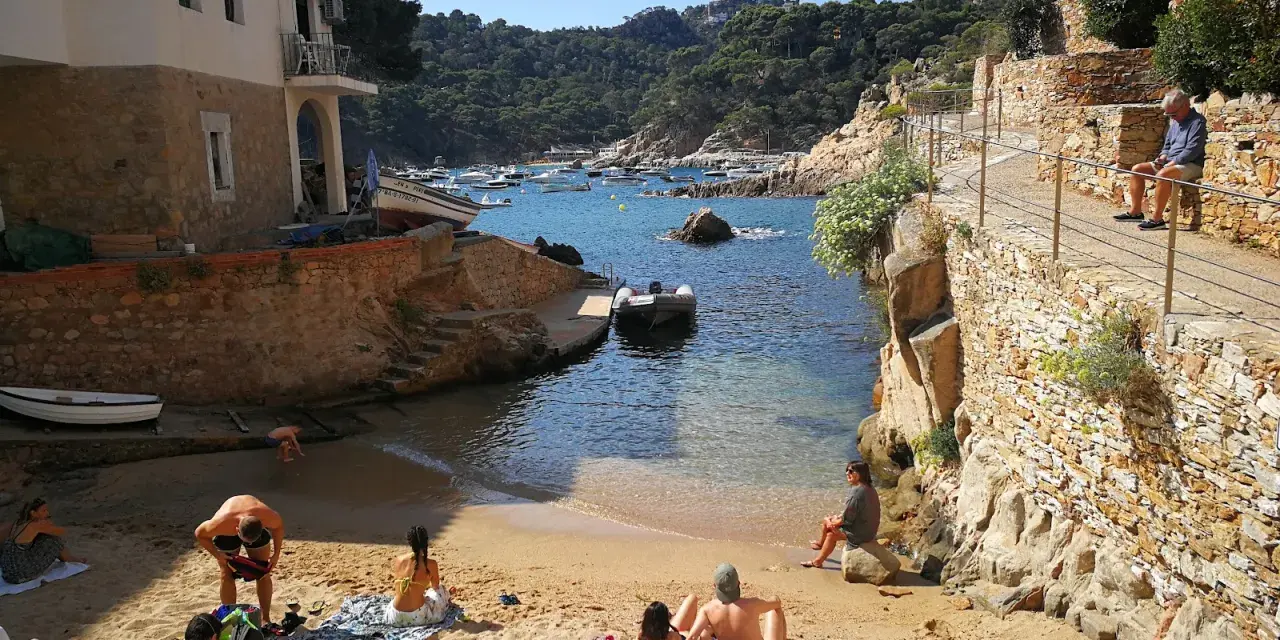 Gente relajándose en la arena de cala Malaret, Begur, con barcos anclados en aguas cristalinas y colinas verdes al fondo.