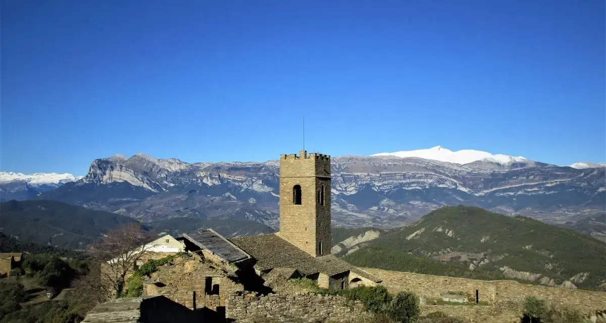 Antiguo muro de roda con torreón de piedra y ruinas, rodeado de montañas nevadas bajo un cielo azul intenso.