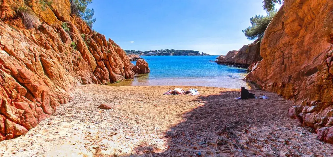 Arena dorada y aguas turquesas en la cala Maset. Dos personas toman el sol tranquilamente en la playa, rodeadas de imponentes rocas rojizas.