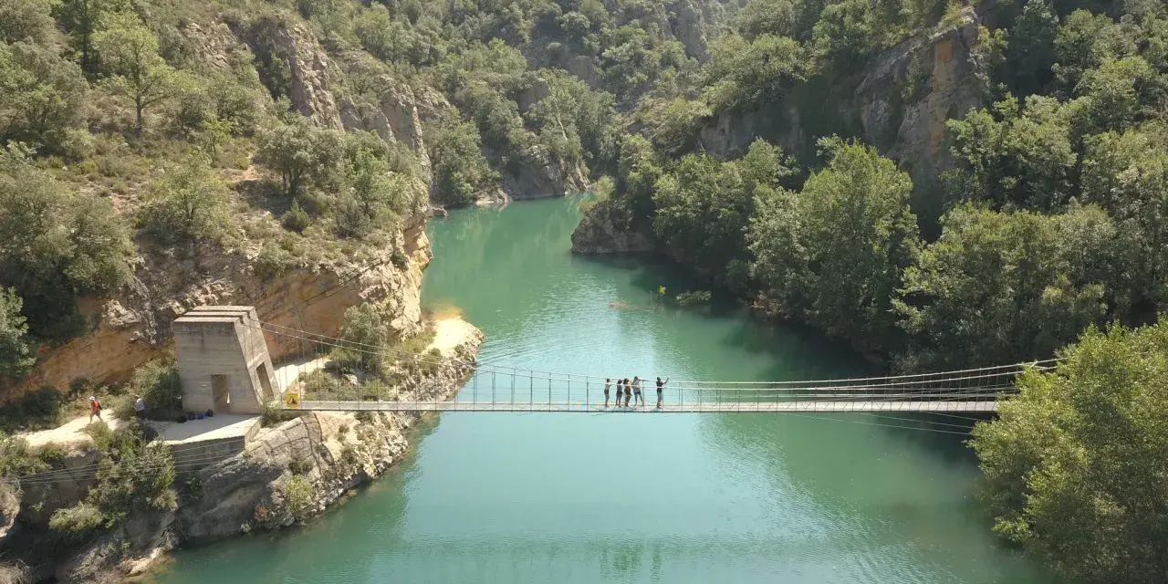 Un puente colgante cruza un río turquesa rodeado de montañas verdes. Un grupo de personas camina por el puente de Montañana.